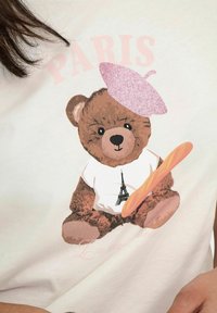 Lysfarvet t-shirt med en bjørn i en lyserød baret, der holder en baguette, samt et Eiffel-tårn grafik på en hvid skjorte.