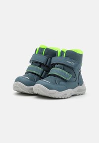 Botas tobilleras sintéticas azules con acentos verde brillante, aseguradas por dos correas de velcro, con una suela de goma texturizada en gris.