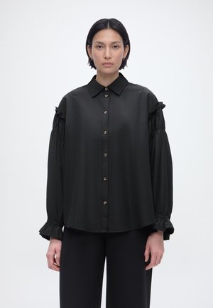 Žena s krátkymi tmavými vlasmi, oblečená v voľnej čiernej blúzke s gombíkmi a volánovými dlhými rukávmi a čiernych nohaviciach, stojaca pred jednoduchým pozadím.