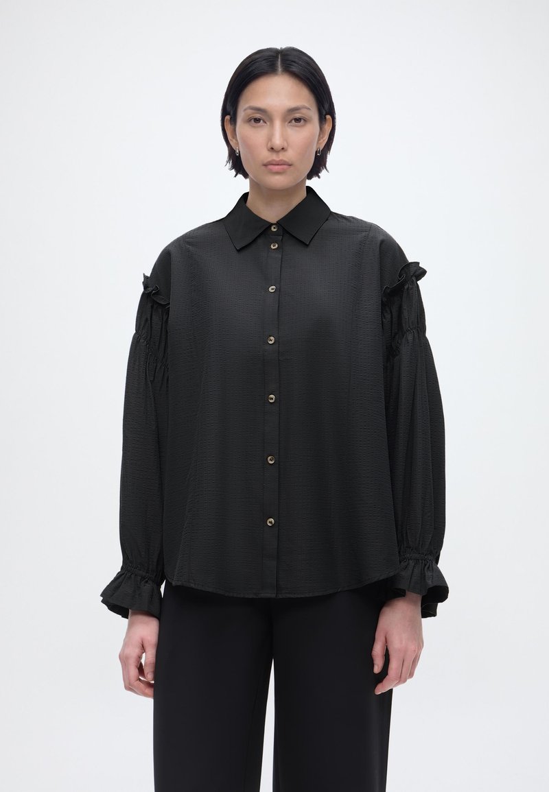 Γυναίκα με κοντά σκούρα μαλλιά που φοράει άνετο μαύρο πουκάμισο με κουμπιά και μακριά φουσκωτά μανίκια, καθώς και μαύρο παντελόνι, στέκεται μπροστά σε απλό φόντο.