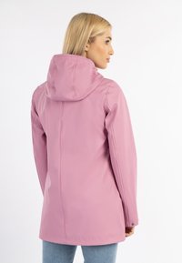 Kvinna med blont hår klädd i en lång rosa regnjacka med huva och blå jeans, vänd bort mot en vit bakgrund.