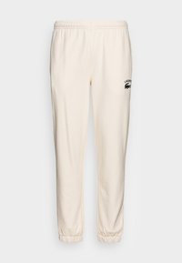 Pantalon de survêtement crème avec une taille élastique, des poignets resserrés et un logo Lacoste noir sur la cuisse gauche. Tissu doux avec une texture lisse.