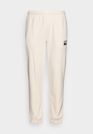 Pantalon de survêtement crème avec une taille élastique, des poignets resserrés et un logo Lacoste noir sur la cuisse gauche. Tissu doux avec une texture lisse.