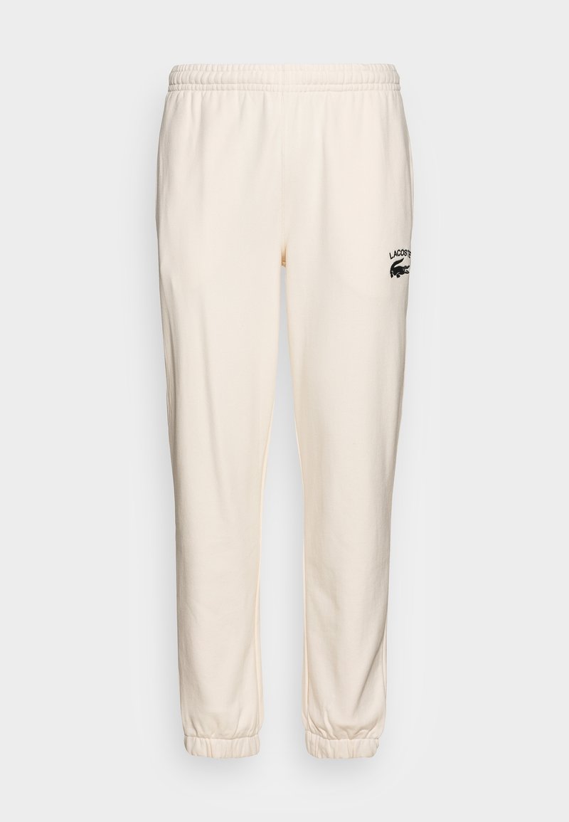 Pantalon de survêtement crème avec une taille élastique, des poignets resserrés et un logo Lacoste noir sur la cuisse gauche. Tissu doux avec une texture lisse.