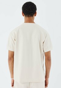 Camisa de manga corta beige con una superficie texturizada, cuello redondo y corte recto. Sin patrones visibles ni detalles de hardware.