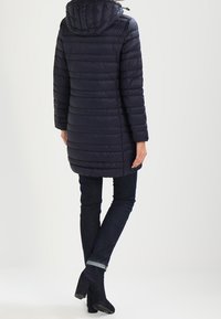 Marineblaue Pufferjacke mit Kapuze, vertikalen Nähten, langen Ärmeln und mittellanger Passform. Kombiniert mit dunklen Jeans und schwarzen Stiefeletten.