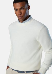 Weißer gestrickter Pullover mit strukturiertem Muster, Rundhalsausschnitt und gerippten Bündchen. Darüber wird ein blau-weiß gestreiftes Hemd getragen.
