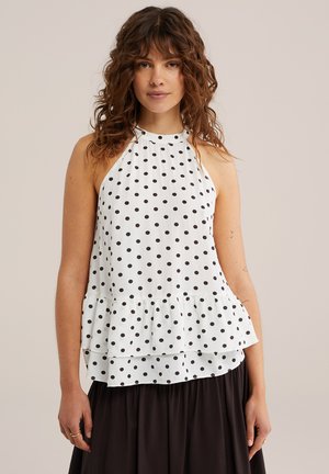 Žena s kučeravými hnedými vlasmi, oblečená v bielom tielkovom top s čiernymi bodkami a čiernu sukňu, stojaca pred jednoliatym pozadím.