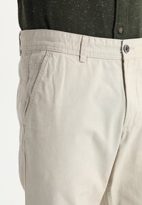 Beige cotton chinos with a relaxed fit, featuring side pockets, a button closure, and subtle texture. Outfit includes a dark green speckled shirt.