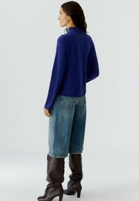 Navy gebreid sweater met hoge kraag, gecombineerd met lichtblauwe wijde jeans en donkere kniehoge laarzen, die een ontspannen silhouet weergeven.