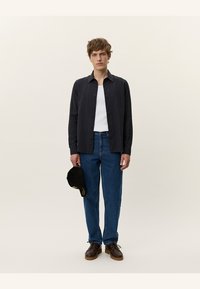Dark, ribbed button-up shirt over a white tank top, paired with blue jeans and brown shoes. Holding a black cap with gold accents.