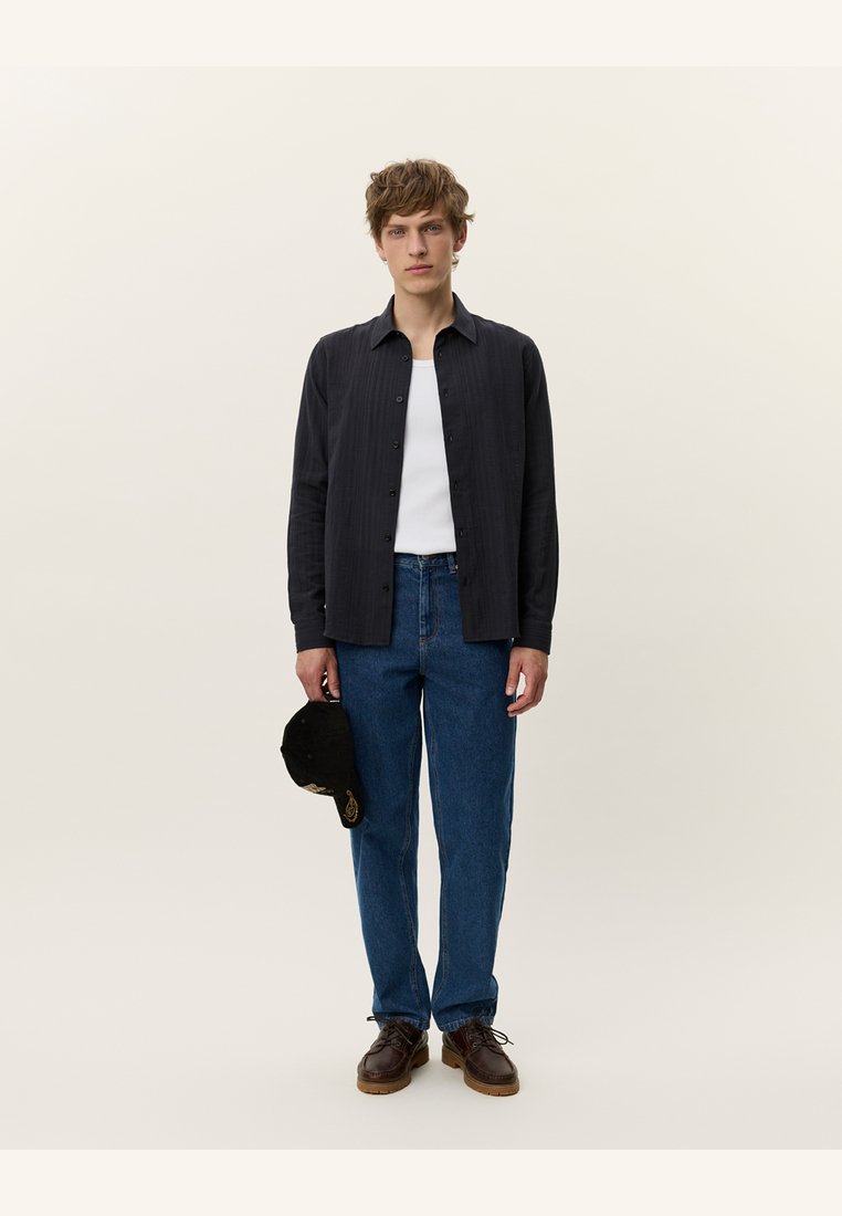 Dark, ribbed button-up shirt over a white tank top, paired with blue jeans and brown shoes. Holding a black cap with gold accents.