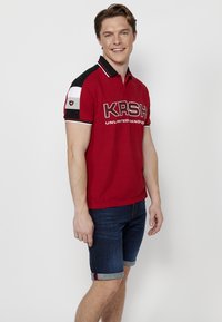 Piros póló fekete kiegészítőkkel és fehér részletekkel az ujjakon, amelyen egy merész "KRSH" logó látható. Sötét farmer sorttal viselve.