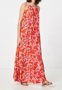 Robe maxi florale rouge avec des fleurs roses et blanches, sans manches, encolure froncée et ourlet à volants. Associée à des sandales plates marron.