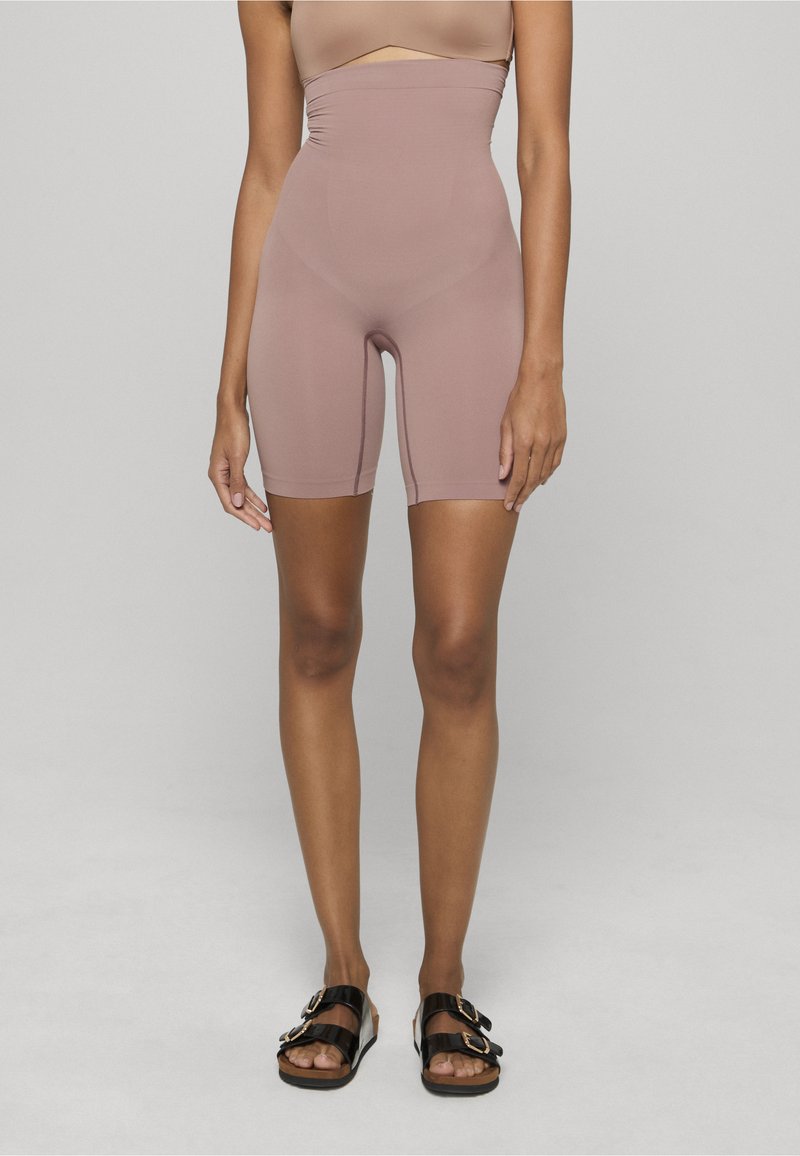 Kobieta ubrana w różowe spodenki rowerowe z wysokim stanem i bezszwowym pasem, w czarnych sandałach na podwójnej klamrze, stoi na jasnoszarym podłodze i tle.