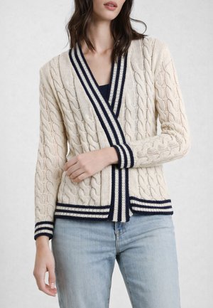 Donna che indossa un cardigan color crema a trecce con bordo a righe blu scuro sopra una maglia scura e jeans azzurri, in piedi davanti a uno sfondo neutro.