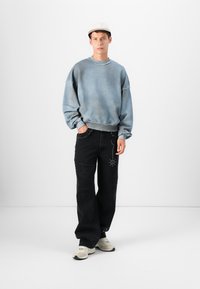 Sweatshirt oversized azul claro com punhos canelados, combinado com calças largas pretas desgastadas com tachas decorativas, usado com ténis bege.