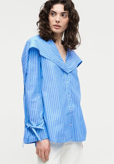 Γυναίκα με καστανά, σγουρά μαλλιά, φοράει μια μπλε και λευκή πουκαμίσα με κάθετες ρίγες, μεγάλο γιακά και μανσέτες με κορδόνια, συνδυασμένη με λευκό παντελόνι.