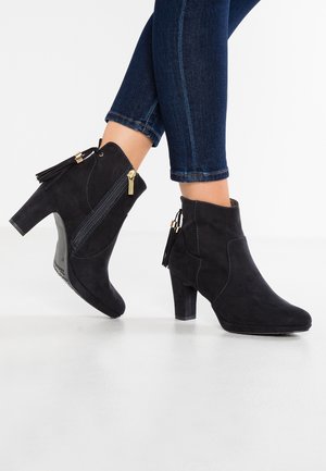 Bottes cheville en daim noir avec talons carrés, fermetures éclair latérales et détails à pampilles, portées avec un jean bleu foncé retroussé.
