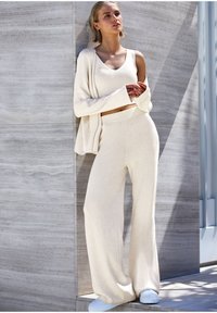 Svetlo béžový rebrovaný pletený crop top a súprava širokých nohavíc, v kombinácii s bielymi teniskami. Modelka sa opiera o textúrovanú sivú stenu.