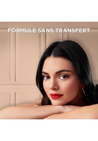 Modèle avec de longs cheveux noirs, portant du rouge à lèvres et des boucles d'oreilles en or. Peau lisse contre un fond beige légèrement texturé.