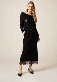 Top negro con escote off-shoulder y detalle plateado; falda larga negra con detalles de encaje en el dobladillo. Combinado con tacones negros de punta afilada.