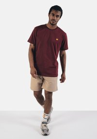Bordeauxfarbenes Baumwoll-T-Shirt mit einem kleinen gelben Elefantenlogo, kombiniert mit beigen Shorts. Das Modell trägt graue Sneakers und gemusterte Socken.
