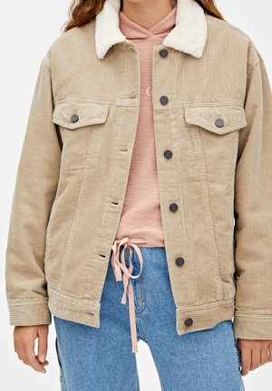 Person iført beige kordjakke med hvid sherpa-krave, pink hættetrøje med snore og blå jeans, vist fra skuldre til hofter.