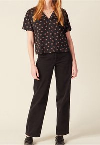 Donna che indossa una blusa nera a maniche corte con scollatura a V a fiori, pantaloni neri larghi e mocassini neri, in piedi davanti a uno sfondo semplice.