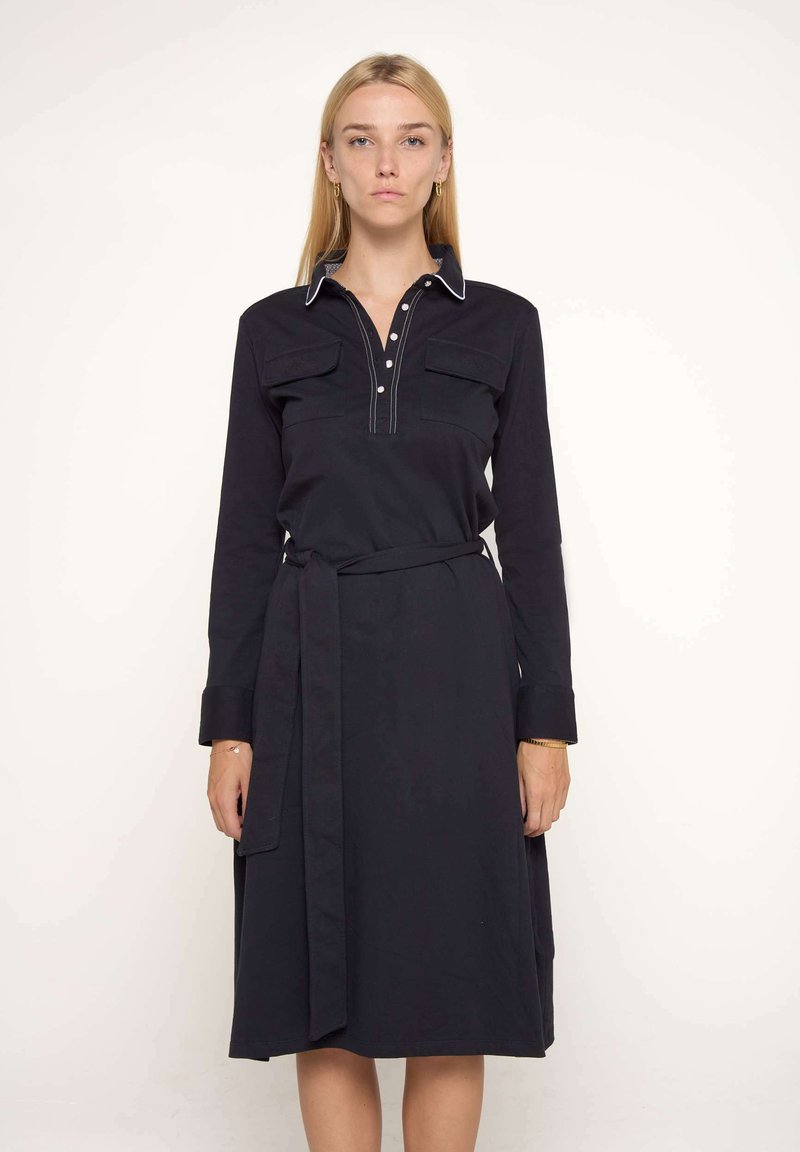 Robe noire à manches longues en tissu doux, dotée d'un plastron à boutons, de deux poches poitrine et d'une ceinture nouée à la taille.