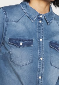 Denimskjorta i ljusblå färg, med spetsig krage och knäppning framtill. Inkluderar två bröstfickor med knappflikar.