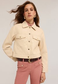 Zkrácená krémová umělá kožená bunda se dvěma klopovými kapsami, zlatými patentkami a špicatým límcem, doplněná o růžové kalhoty a tmavý opasek.