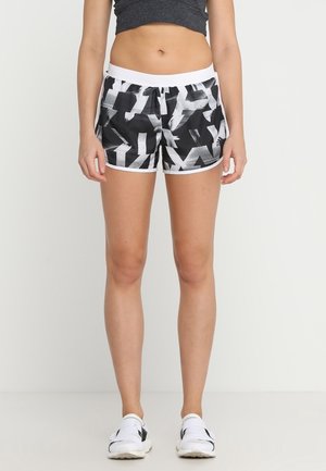 Kobieta stoi, ubrana w czarno-białe sportowe szorty w abstrakcyjny wzór, ciemnoszary krótki top i białe buty do biegania.