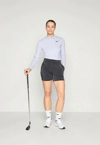 Egy nő világos, hosszú ujjú felsőt, fekete sortot, fehér zoknit és sportcipőt visel, egyik kezével zsebébe, a másikkal pedig egy golfütőt tart.