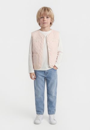 Blond chlapec má na sebe svetloružovú prešívanú vestu cez bielu košeľu s dlhým rukávom, svetlomodré džínsy a biele tenisky, stojí na bielom pozadí.