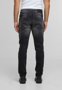Mann iført falmede sorte jeans, sorte sko og en hvid ærmeløs skjorte, som står vendt væk med en ensfarvet grå baggrund.