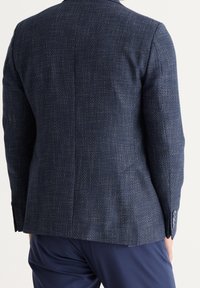 Muž v tmavě modrém texturovaném sáčku a námořnicky modrých kalhotách, zobrazený zezadu na jednoduchém světlém pozadí.