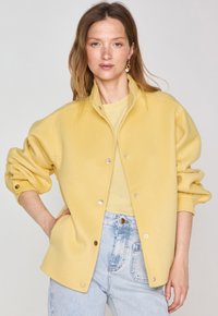 Yellow fleece jacket with a collared neckline and snap buttons, featuring elastic cuffs and a relaxed fit, worn over a light shirt.