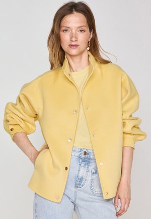 Giacca in pile gialla con colletto e bottoni a pressione, dotata di polsini elasticizzati e vestibilità comoda, indossata sopra una camicia leggera.