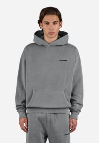 Sudadera gris de tela suave, con bolsillo canguro, puños acanalados y un pequeño logo en el pecho. Diseño minimalista.