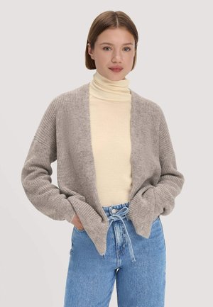 Junge Frau mit kurzen braunen Haaren, die einen beigen Rollkragenpullover, eine hellgraue Strickjacke und hellblaue, hoch taillierte Jeans mit gebundenem Gürtel trägt.