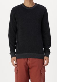 Maglione in maglia nera con texture, collo rotondo, polsini e vita a coste, indossato sopra pantaloni cargo rossastri.