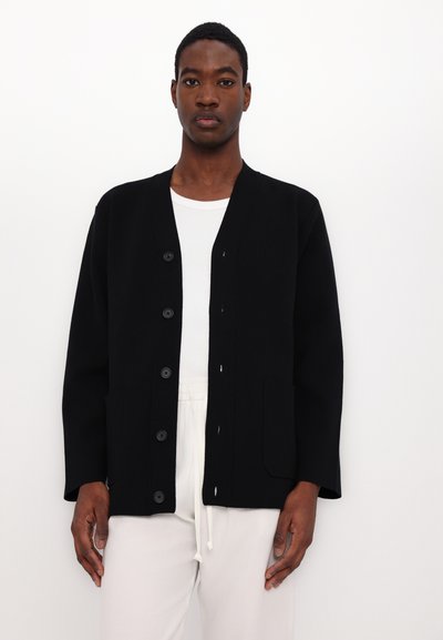 Black cardigan made of textured fabric, featuring a V-neck, six buttons, and two front pockets. Worn over a white T-shirt.