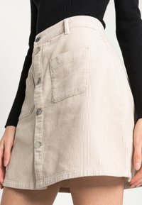 Beige kordskjol med A-linjeform, framknäppning och två sidfickor. Ribbad textur och nederkant som slutar ovanför knäet.