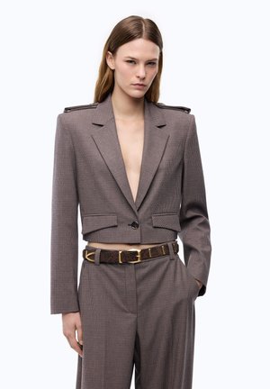 Joven mujer que lleva una blazer corta a cuadros a medida y pantalones de cintura alta a juego con un cinturón marrón, de pie con una mano en el bolsillo.