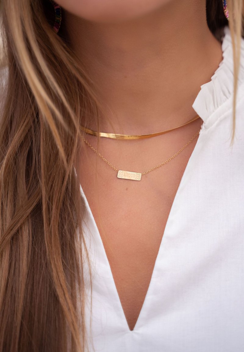 Choker en or et collier avec pendentif superposés sur un col blanc. Le choker a une texture lisse ; le pendentif est rectangulaire avec des détails gravés.