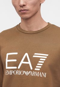 Νέος άνδρας φοράει καφέ μπλουζάκι με λευκό λογότυπο EA7 Emporio Armani μπροστά.