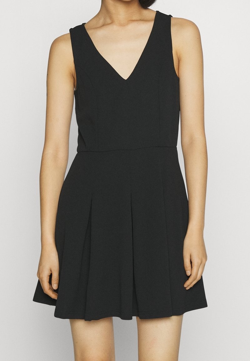 Czarna sukienka bez rękawów z głębokim dekoltem w kształcie litery V i plisowanym dołem, wykonana z gładkiego materiału. Charakteryzuje się minimalistycznym stylem i czystymi liniami.