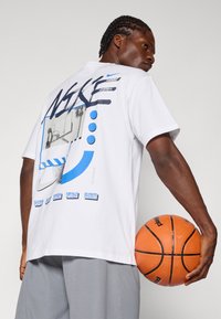 Λευκό T-shirt με μπλε γραφιστικό σχέδιο και κείμενο στο πίσω μέρος, συνδυασμένο με γκρι σορτσάκι. Το άτομο κρατάει ένα μπάσκετ.