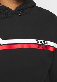 Felpa nera con cappuccio, nella zona del petto presenta strisce orizzontali bianche e rosse e il testo "TOMMY JEANS" sul lato destro, sotto il colletto.